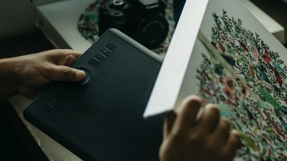Grafik tablet tutan kişi, kamera yanındaki çiçek desenli ürün kutusunu inceliyor.