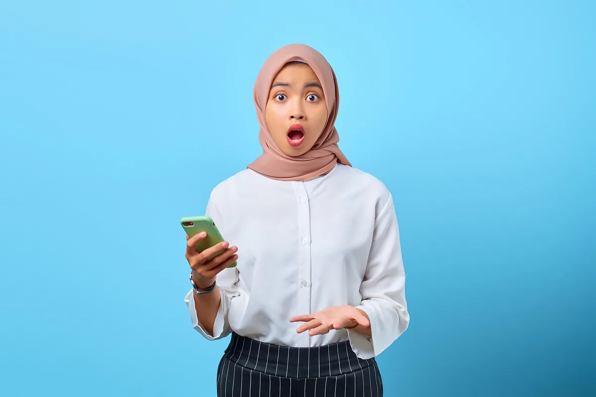بانویی با روسری صورتی پودری که در برابر پسزمینه آبی با شگفتی به گوشی خود نگاه میکند