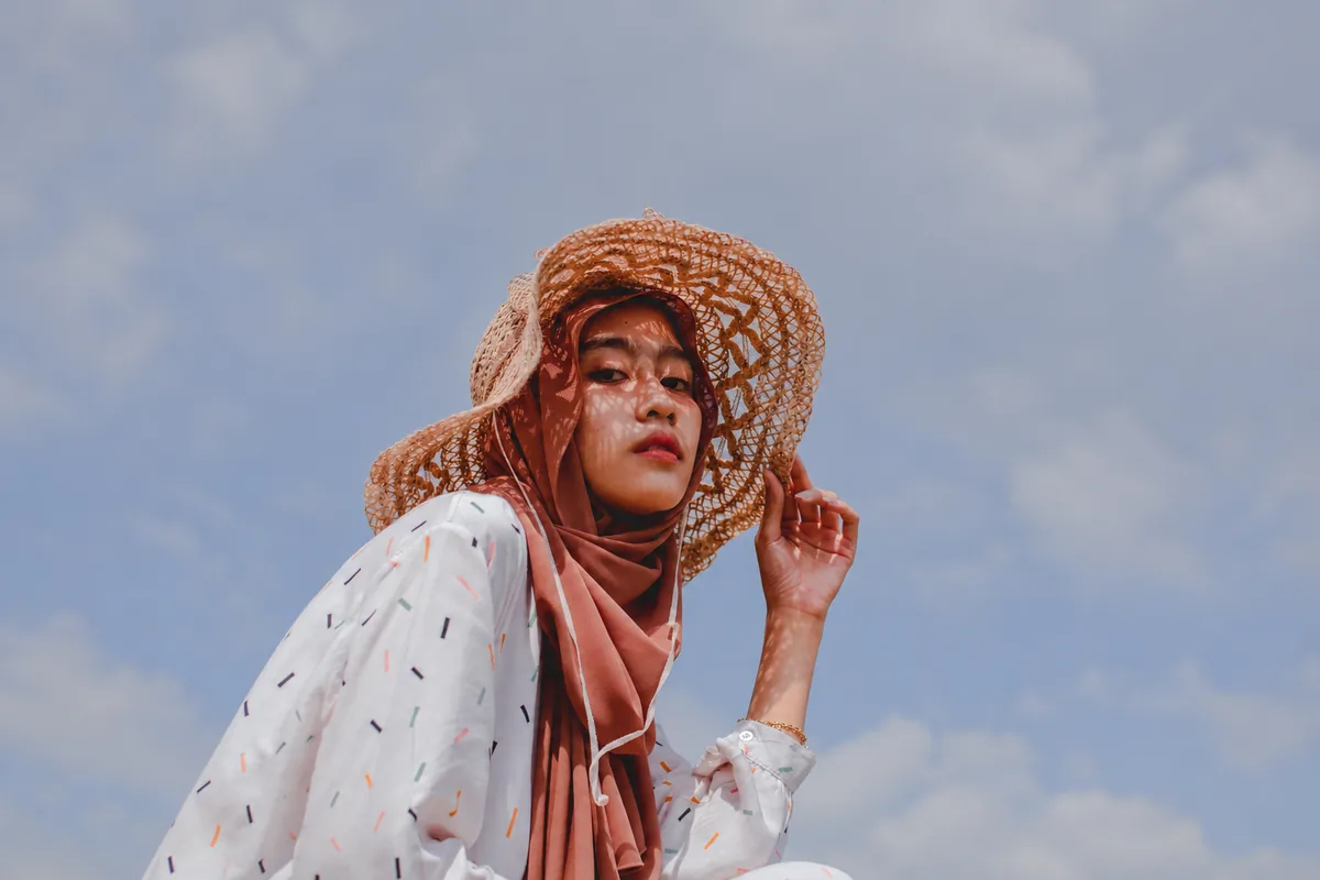 بانویی با کلاه بافتنی و روسری صورتی پودری که در برابر آسمان ابری ژست گرفته است